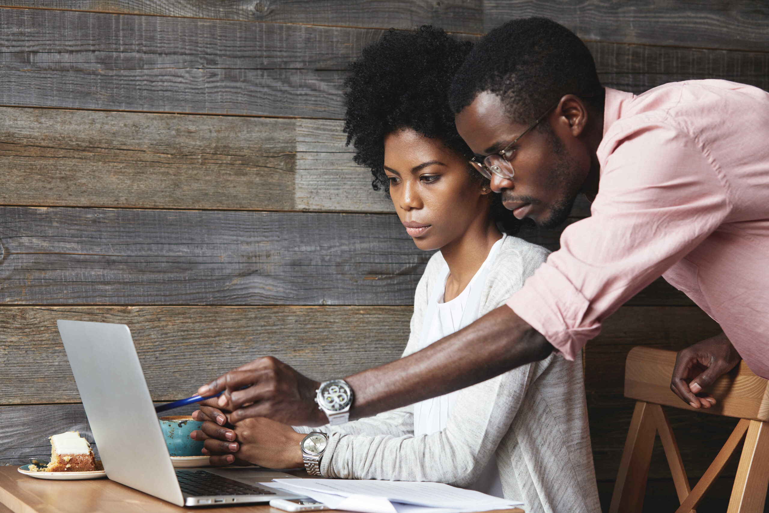 People and education concept. Young African teacher in glasses explaining new material on economics to his beautiful female student, pointing at laptop screen with pen while having lesson at cafe